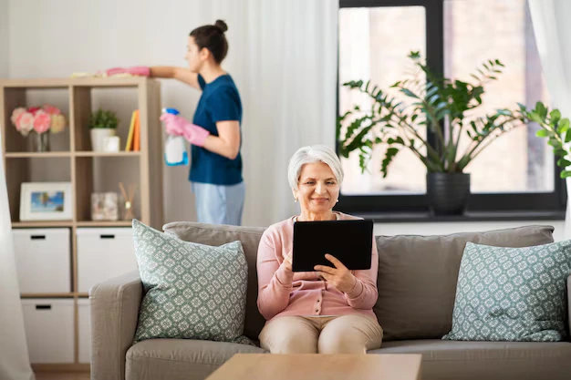 Older woman relaxing while woman professional cleaner dusting at home during senior-friendly house cleaning services in Ajax